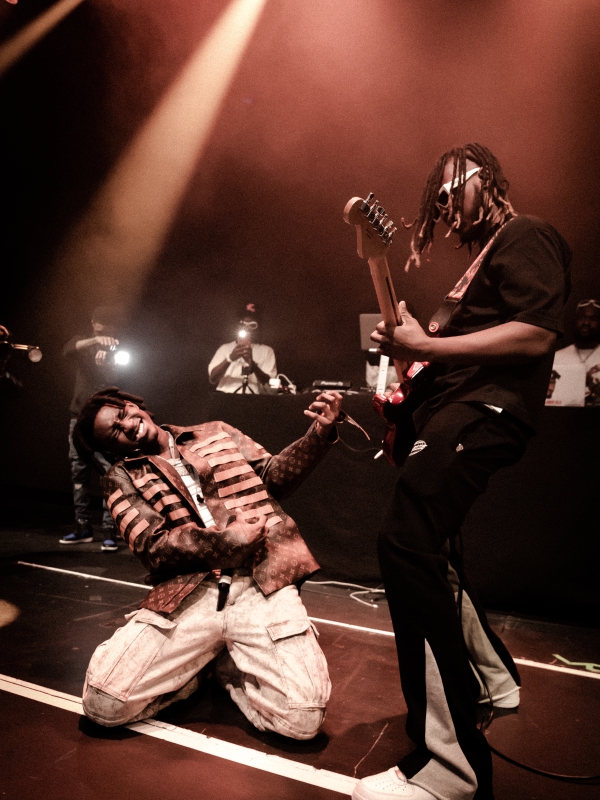 Dynamic stage shot of a Shallipopi kneeling on stage while a guitarist performs beside him under a warm spotlight. — concert photography London by Lamss Media